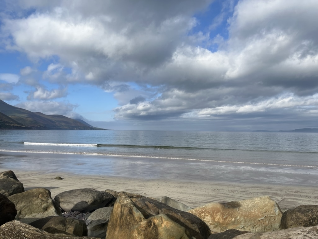 A wide, peaceful view of Rossbeigh Strand on the Ring of Kerry, showing calm waves, sandy beach, rocky foreground, and mountains under a cloudy blue sky.