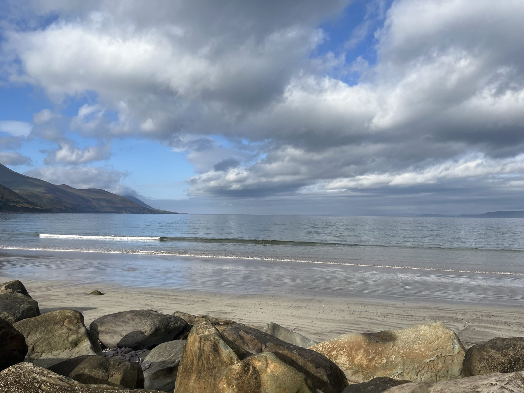 A wide, peaceful view of Rossbeigh Strand on the Ring of Kerry, showing calm waves, sandy beach, rocky foreground, and mountains under a cloudy blue sky.