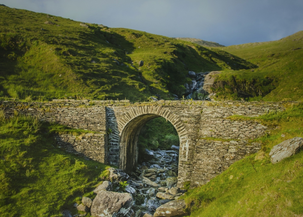 Stone arch bridge over a rocky stream in Healy Pass, surrounded by green mountains and rugged Irish landscape.