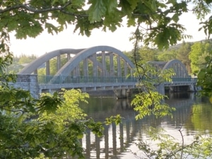 Arched bridge in Kenmare reflected in calm river water, surrounded by green foliage on a sunny day.