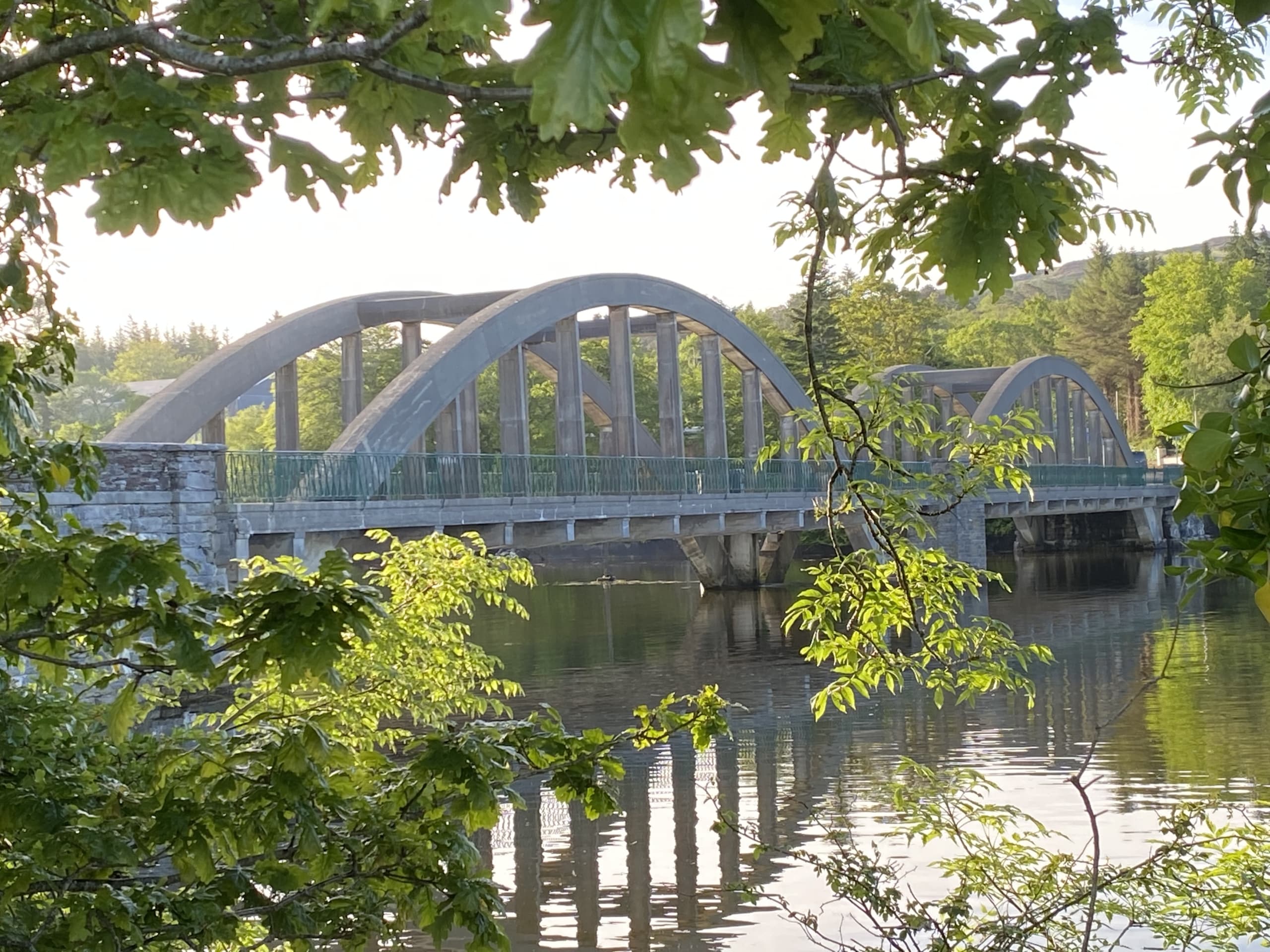 Arched bridge in Kenmare reflected in calm river water, surrounded by green foliage on a sunny day.