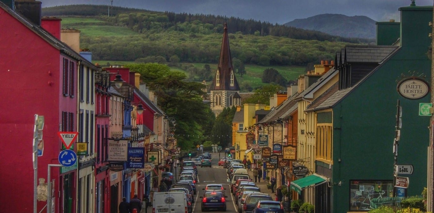 A colourful street view of Kenmare Town with busy shopfronts, parked cars, and Holy Cross Church rising in the background against green hills.