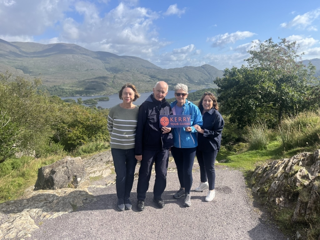 A group of four guests smiling and holding a Kerry Tour Guides sign with the mountains and lakes of Killarney National Park in the background.