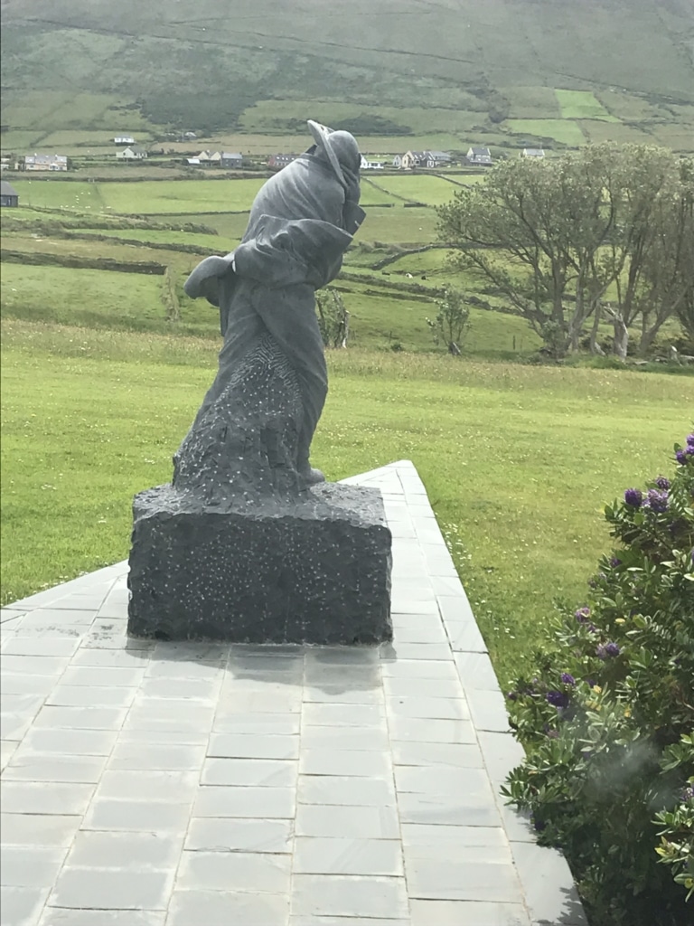 Stone sculpture called The Islandman at the Blasket Centre in Dunquin, set against green fields and hills on the Dingle Peninsula.