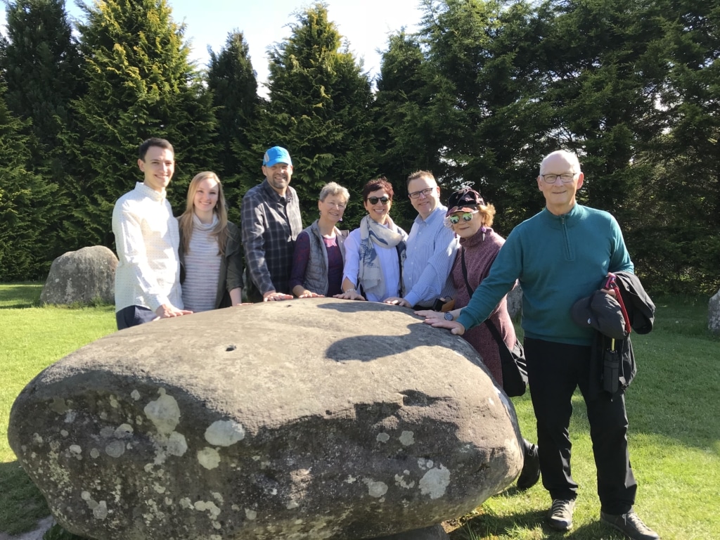Group of visitors standing around a large stone at an ancient stone circle, smiling in a sunny outdoor setting surrounded by trees.